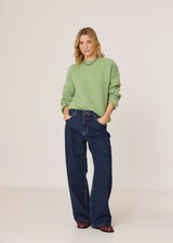 A woman modeling a light green knit crewneck sweater and dark blue wide-leg jeans against a plain light beige studio background.