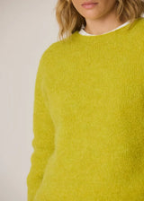 A close-up of a person wearing a fuzzy, textured knit sweater in a bright chartreuse yellow-green color, layered over a white crewneck shirt, against a plain light background.
