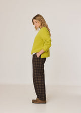 A woman stands in profile wearing a bright yellow-green knit sweater, dark brown and black plaid trousers, and brown shoes against a plain light gray studio background.