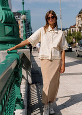A woman stands on a city bridge wearing a cream short-sleeve button-up shirt with lace trim and a beige midi skirt, paired with white sneakers. She is leaning against a green metal railing, with buildings and a street visible in the background.