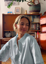 A woman with blonde hair wearing a light blue short-sleeve button-up shirt with lace trim, standing in a cozy room with a wooden bookshelf and a double bass in the background.