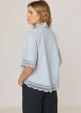 A woman shown from the back wearing a light blue short-sleeve button-up shirt with scalloped lace trim at the cuffs and hem, paired with dark pants, against a plain light background.