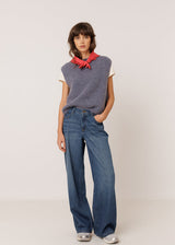 A model stands against a light beige studio background wearing a gray sleeveless knit vest over a white t-shirt with a red neck scarf, paired with wide-leg blue jeans and silver sneakers.