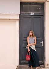 A woman stands in front of a dark charcoal door, wearing a gray sleeveless top, a long navy skirt, and holding a red woven handbag.