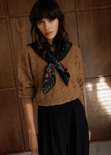 A woman wearing a brown cable-knit cropped sweater, a black patterned scarf tied around her neck, and a black skirt, standing against a dark wood-paneled wall.