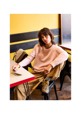 A woman sits on a black metal chair in a cafe setting, wearing a fuzzy light pink crewneck sweater, beige trousers, and yellow sneakers. The background features a yellow wall with black horizontal panels.